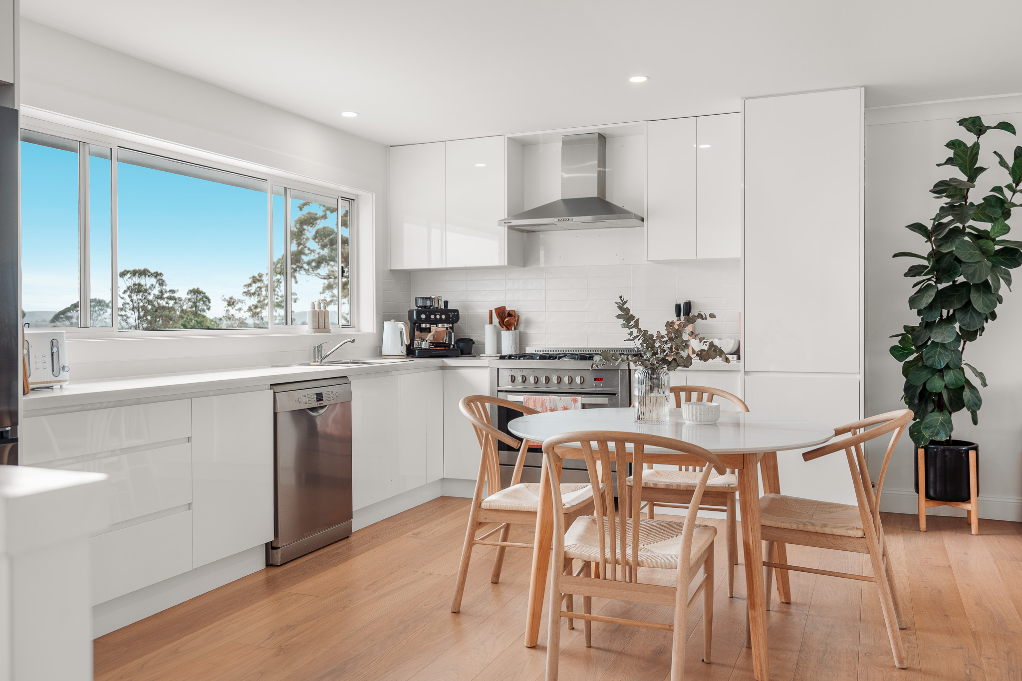 A bright, modern kitchen and dining area in a Bomaderry home built by Porter Building Group, featuring minimalist white cabinetry, stainless steel appliances, and warm timber flooring.